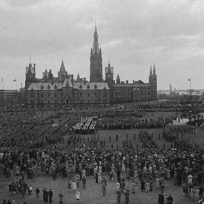 Photographie en noir et blanc – Des milliers de personnes rassemblées près des édifices du Parlement à Ottawa. Une tribune est dressée au milieu de la photo, à droite. Plusieurs véhicules sont visibles dans les environs.