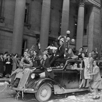 Photographie en noir et blanc – Une voiture roule dans la rue sous des pluies de confettis. Les personnes à l’intérieur et sur son toit manifestent leur joie. Une foule envahit la rue en face d’un gros édifice en béton.