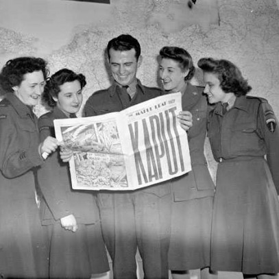 Photographie en noir et blanc – Un homme tient un journal ouvert à une page centrale; la une est couverte de grosses lettres indiquant « KAPUT ». Quatre femmes l’entourent, lisant le journal avec lui.