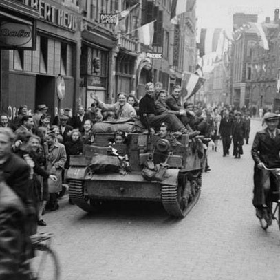 Photographie en noir et blanc – Scène de rue. Des drapeaux néerlandais sont partout, flottant ou brandis. La foule se masse dans les rues et les trottoirs, à pied et en vélo. Un char roule au centre, et des civils y sont montés.