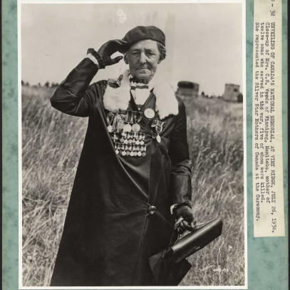 Photographie en noir et blanc, bordée de vert. Une femme âgée portant un béret salue la caméra de la main droite. Elle porte un long manteau noir garni de fourrure et orné de médailles, dont certaines sont miniatures.