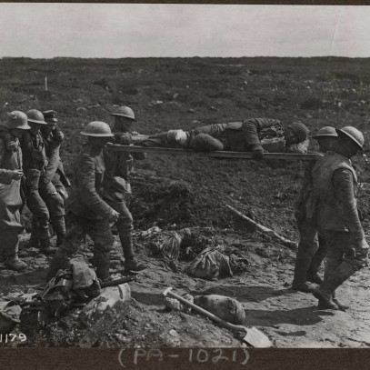 Photographie en noir et blanc – Un champ de bataille boueux. Quatre hommes en uniforme militaire et portant des casques transportent sur leurs épaules un homme allongé sur une civière. Trois hommes les suivent, celui au centre soutenu par les deux autres.