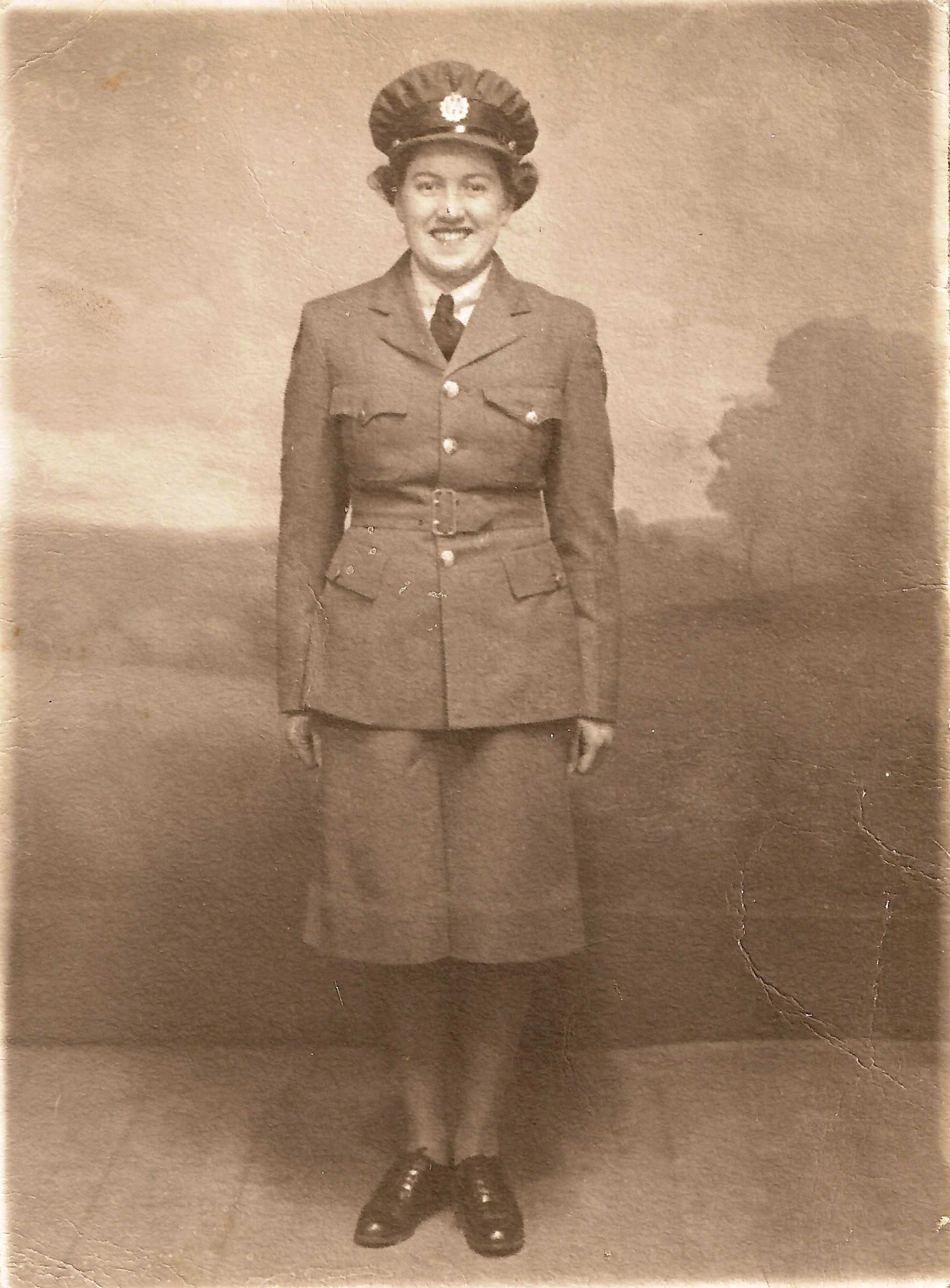 Photographie en tons sépia – Une jeune femme vêtue de son uniforme militaire complet, qui comporte une jupe. Elle est souriante, au garde-à-vous.