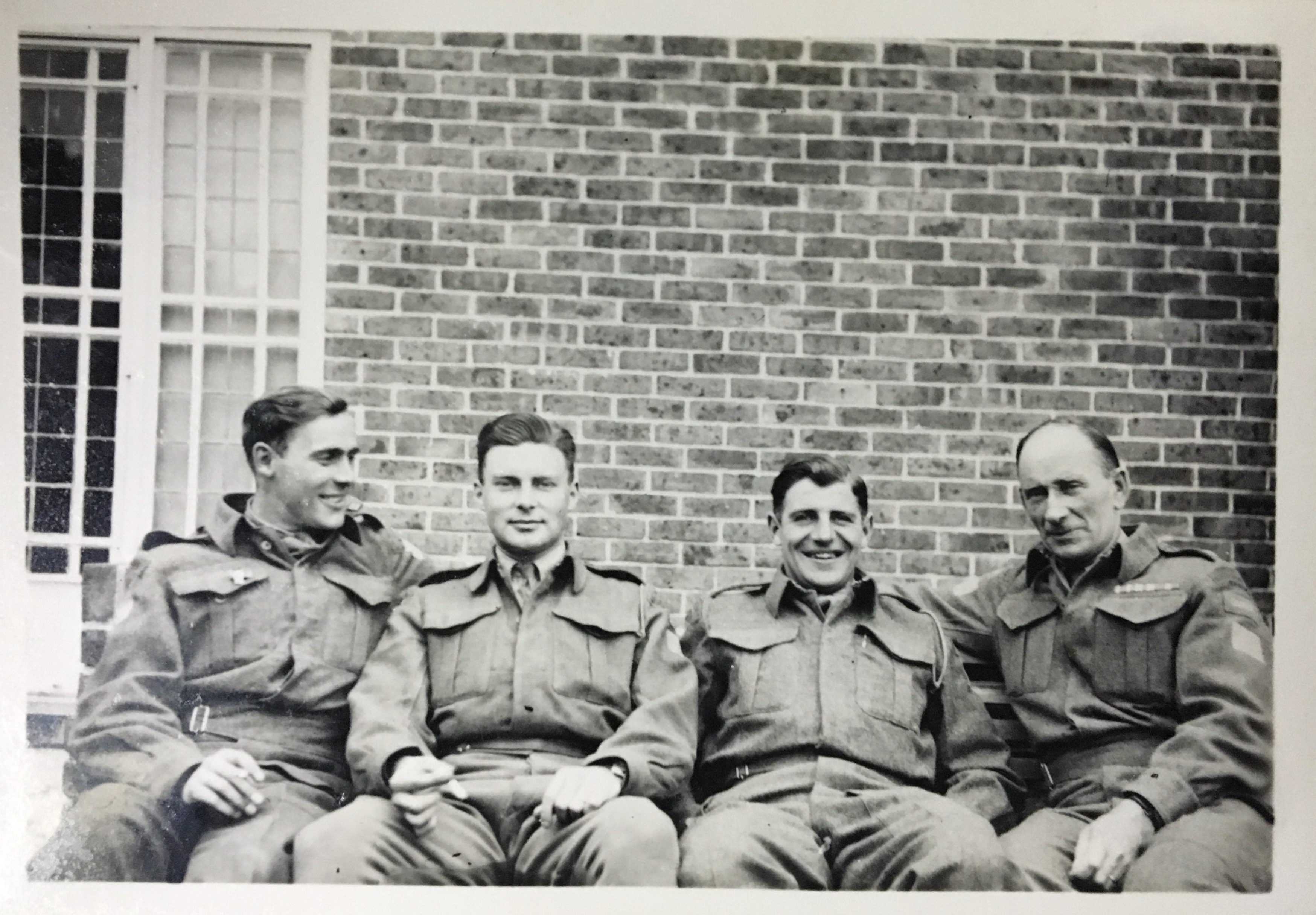 Photographie en noir et blanc – Archie, complètement à droite, est assis sur un banc avec trois hommes beaucoup plus jeunes. Tous sont en uniforme. Ils sourient.