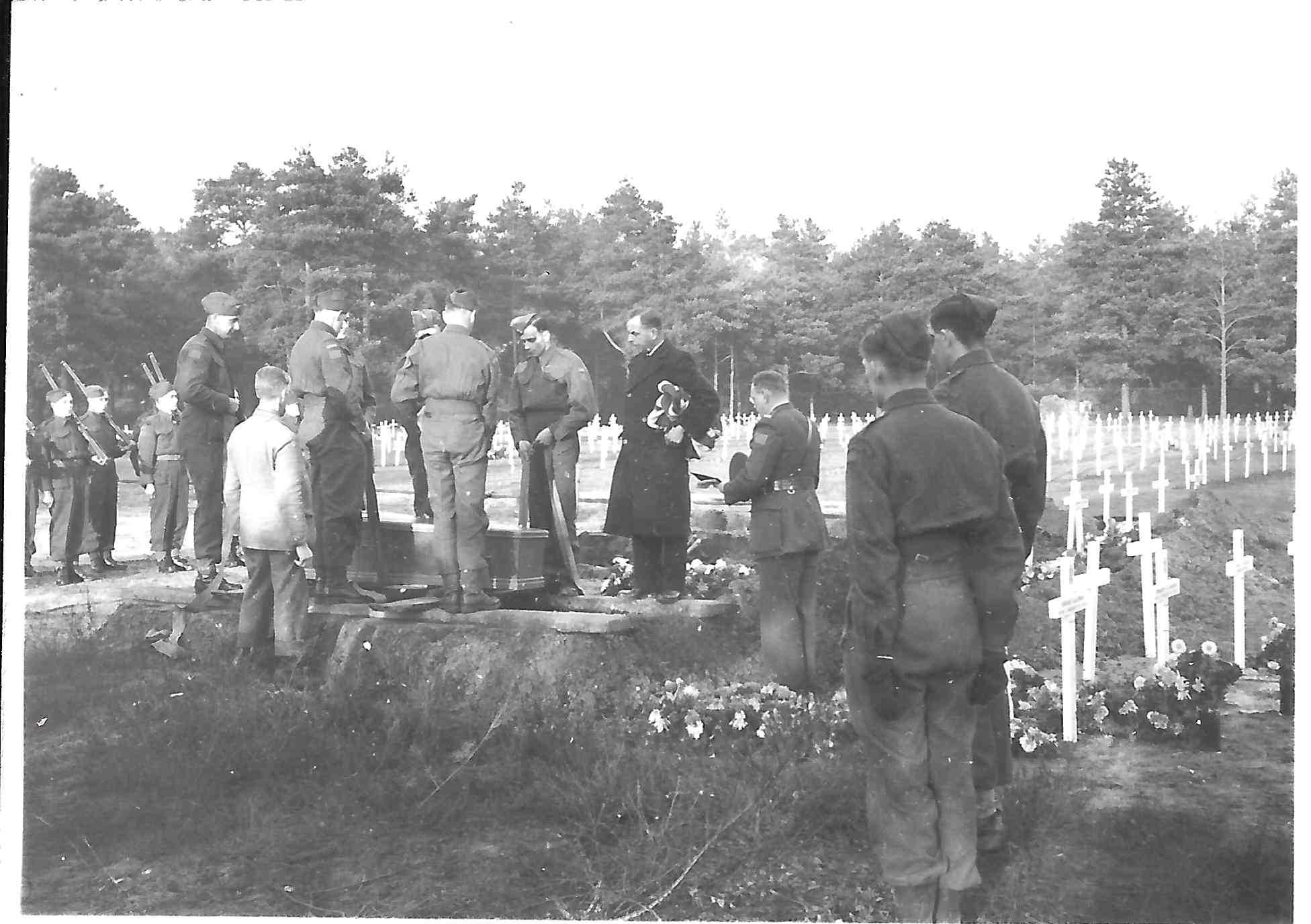Photographie en noir et blanc – Des hommes dans divers uniformes portent en terre un cercueil dans un champ de croix blanches. Un aumônier militaire se trouve devant la tombe.