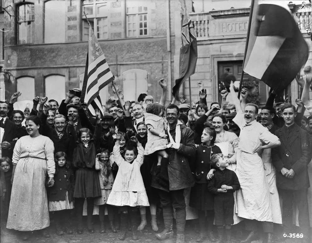Photographie en noir et blanc – Les Canadiens à Valenciennes au sein d’une foule de citoyens français les acclamant. Divers drapeaux de pays alliés sont agités. Tous, hommes, femmes et enfants, sont joyeux.