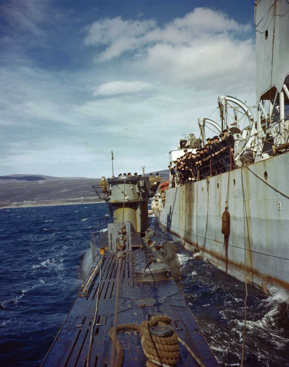 Image en couleur – On aperçoit le bord d’un immense navire, où des marins canadiens s’appuient sur la rambarde. Un sous-marin émergeant à peine de l’eau est fixé au navire au moyen de cordes; des militaires allemands se trouvent dessus.