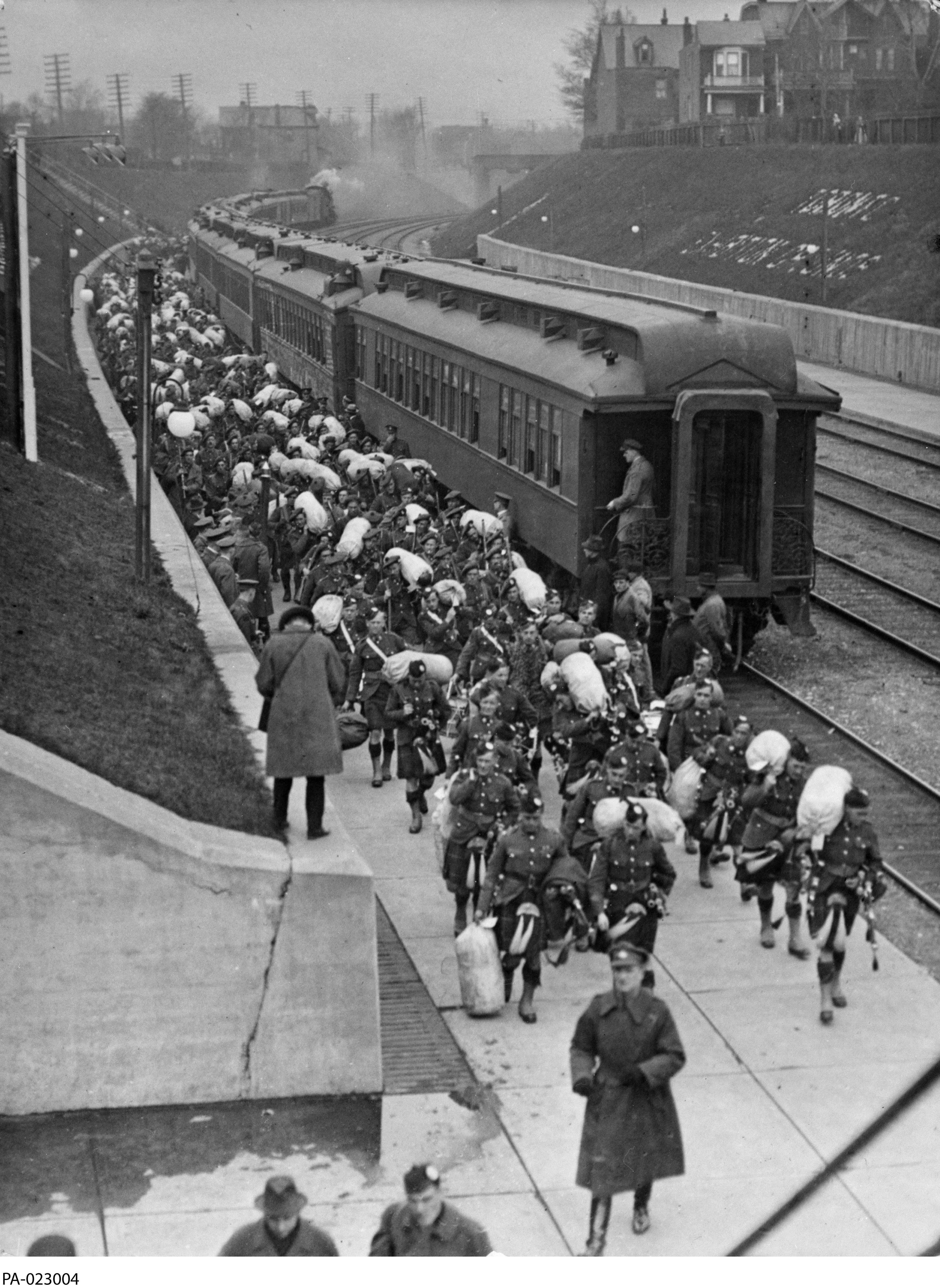 Photographie en noir et blanc – Des centaines de militaires portant un kilt marchent près du train dont ils ont débarqué. La voie ferrée se trouve entre des collines de verdure délimitées par un mur en ciment; un journaliste prend des notes ou une photo.