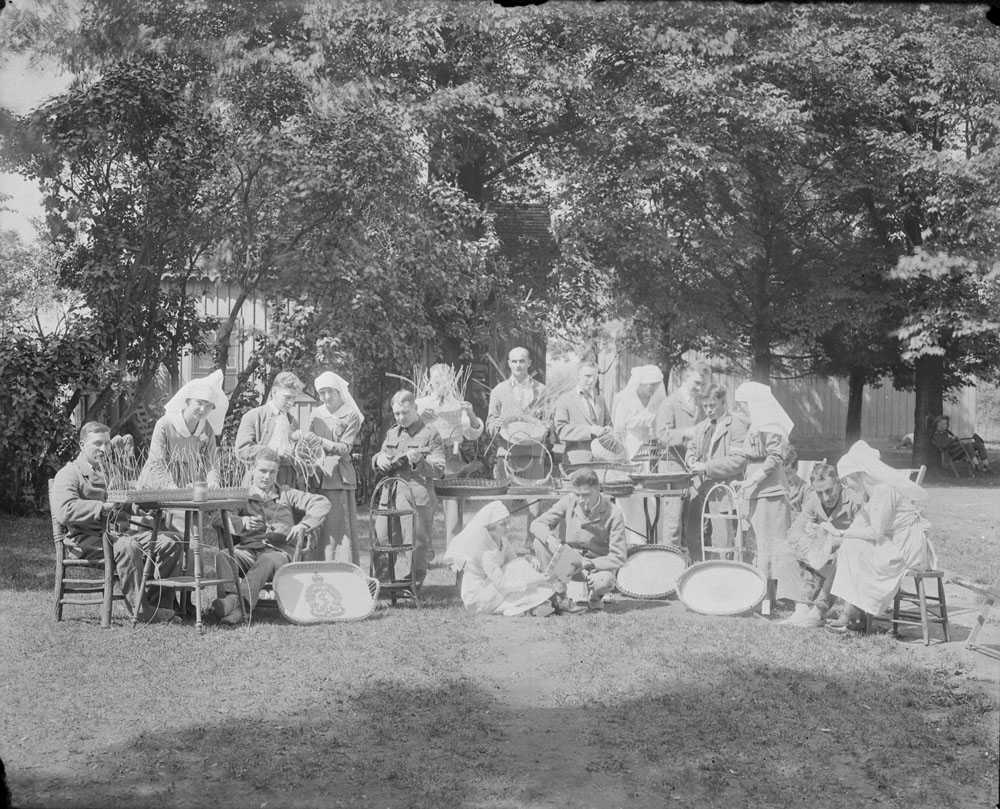 Photographie en noir et blanc – Des infirmières et des soldats sont assis sur des chaises ou sur le sol près d’un grand arbre. Ils sont en train de tisser des paniers.