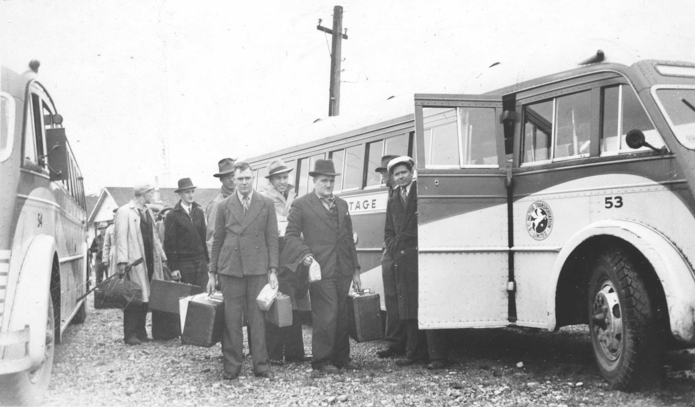 Photographie en noir et blanc – Des hommes bien vêtus rassemblés devant un gros autobus, tenant une valise et attendant au comptoir. La plupart regardent l’objectif de la caméra. Un deuxième autobus se trouve de l’autre côté.