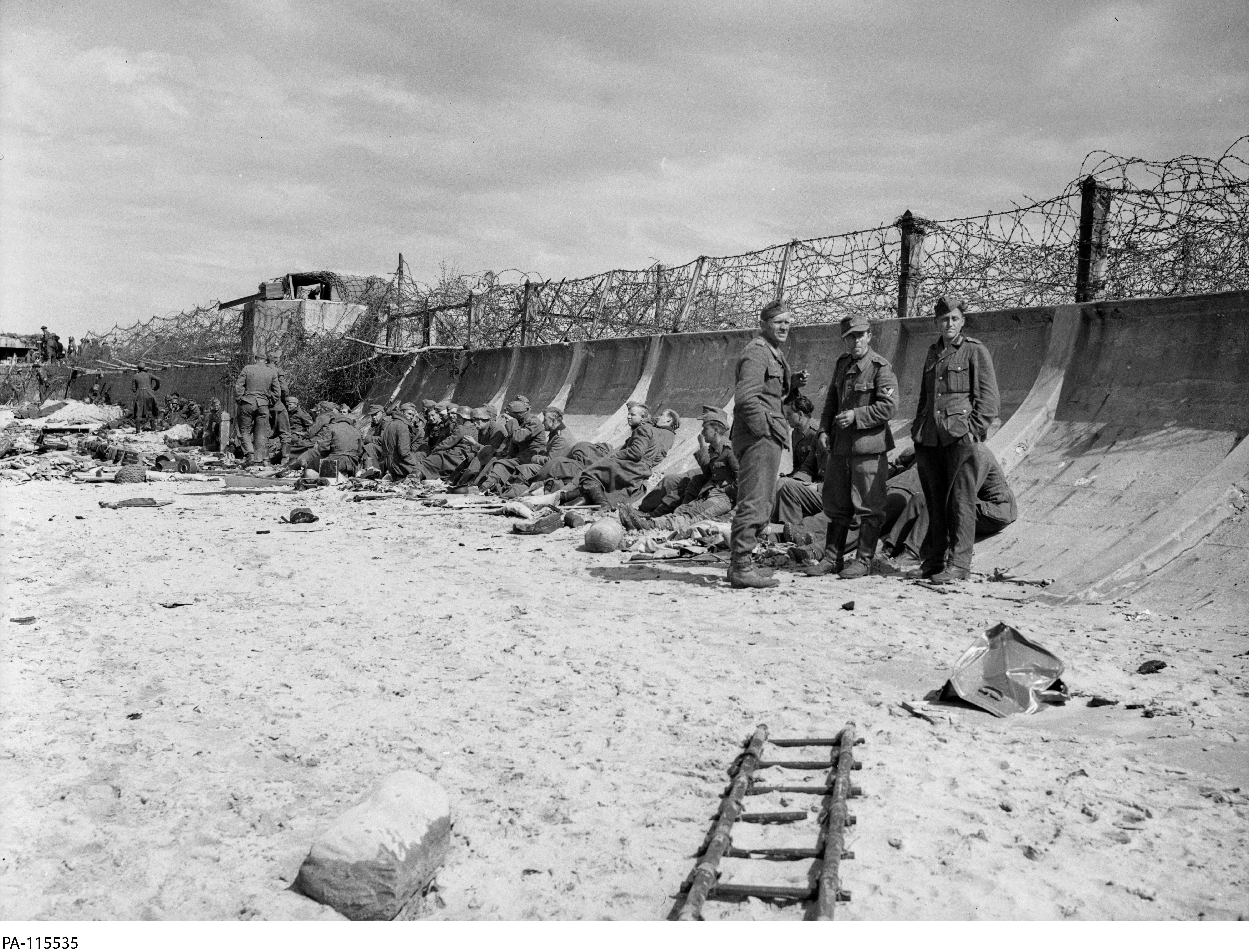 Photographie en noir et blanc – Une plage sablonneuse et une digue élevée, courbée et surmontée d'épais fils barbelés. Un nid de mitrailleuses est visible un peu plus loin sur la plage. Des prisonniers allemands sont assis contre la digue.