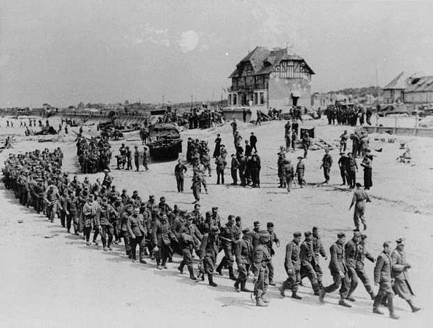 Photographie en noir et blanc – Une longue colonne de soldats allemands avançant sur une plage sablonneuse, sous le regard de militaires canadiens. Des obstacles de plage et des véhicules militaires sont visibles. La Maison du Canada surplombe la scène.