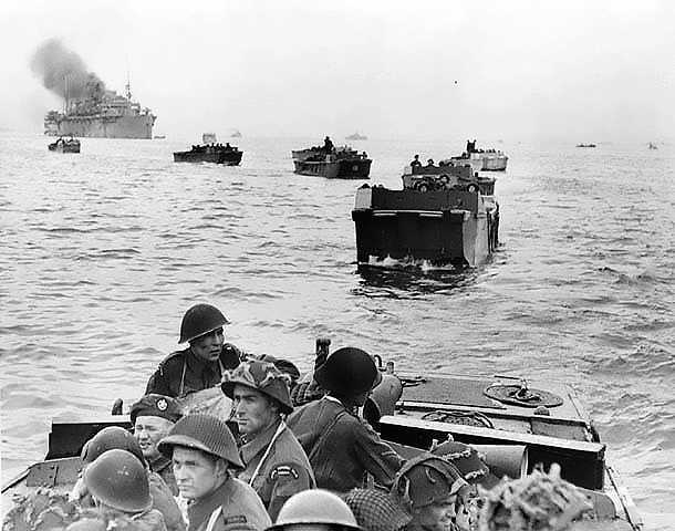 Photographie en noir et blanc – Une série de péniches de débarquement s’éloigne d’un immense navire. Des soldats contemplent la haute mer.