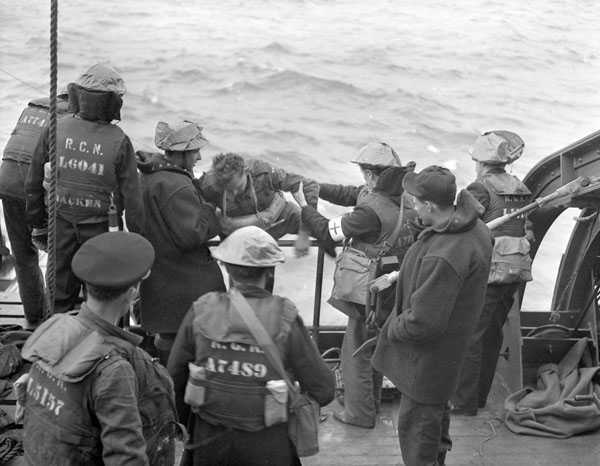 Photographie en noir et blanc– L’extrémité d’un bateau dans une mer agitée. Un soldat est tiré sur le pont par la Marine royale du Canada. La plupart portent des vestes de sauvetage. On reconnaît le Corps de santé royal aux brassards avec une croix rouge.