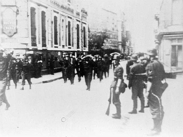 Photographie en noir et blanc – Des soldats canadiens marchent à travers les rues de Dieppe, les mains derrière la tête. Des soldats allemands armés, sur le côté, les surveillent.