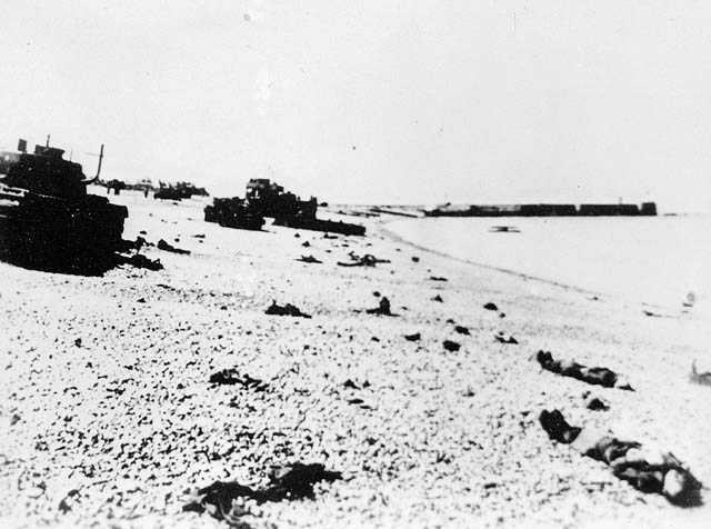 Photographie en noir et blanc – Un quai est visible au loin. Des chars d’assaut sont abandonnés et des corps reposent sur la plage rocheuse.