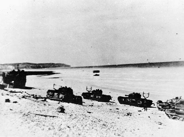 Photographie en noir et blanc – Quatre chars d’assaut et une péniche de débarquement sont abandonnés sur les plages rocheuses de Dieppe. Au loin, on aperçoit de l’équipement militaire reposant dans la mer à marée basse.