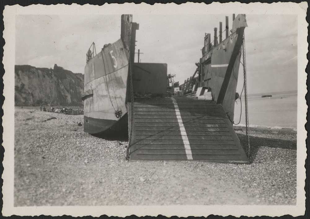Photographie en noir et blanc – Une barge de débarquement de chars d’assaut sur une plage de galets à Dieppe. On aperçoit au loin les falaises blanches emblématiques de la région. La péniche se trouve entièrement sur la plage, et sa plateforme d’embarquem