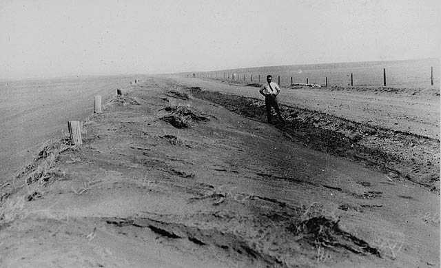 Photographie en noir et blanc – Un homme est debout en bordure de la route longeant des champs. Même si la photo est en noir et blanc, il est facile de dire à quel point le sol est sec. L’air est rempli de poussière.