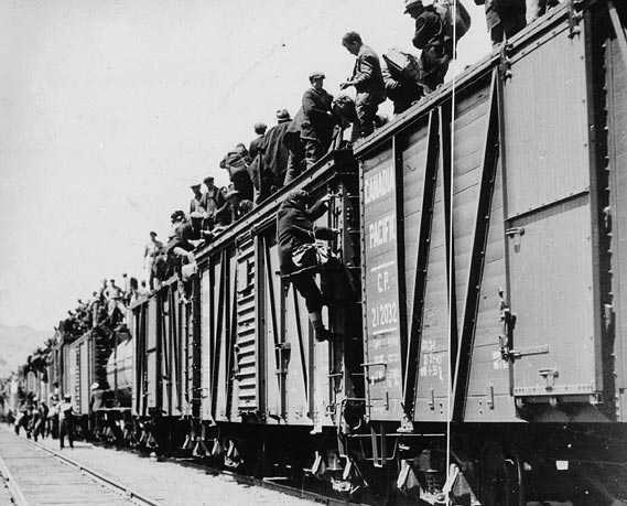 Photographie en noir et blanc – Des wagons avancent sur une voie ferrée. Des hommes y montent et se rassemblent sur le toit. On en comptent des centaines.
