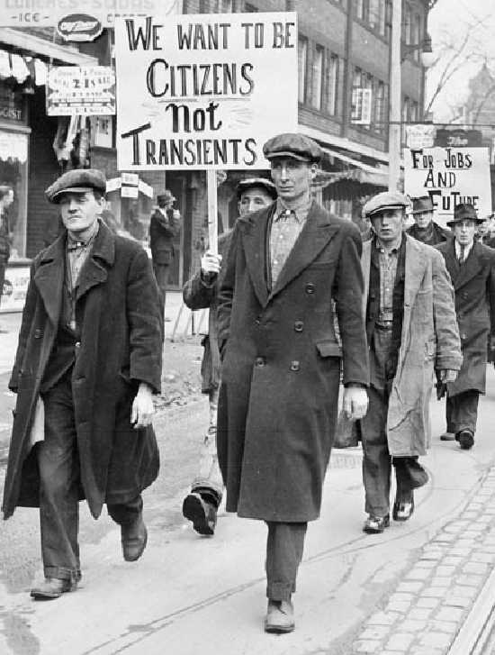Photographie en noir et blanc – Des hommes vêtus de longs manteaux défilent dans une rue de la ville. Plusieurs tiennent des pancartes.