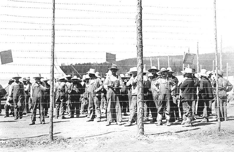 Photographie en noir et blanc – De nombreux hommes vêtus d’une salopette et d’un chapeau à large bord se trouvent derrière une haute clôture de barbelés. Ils ne font rien. Une tente est visible à l’arrière-plan.
