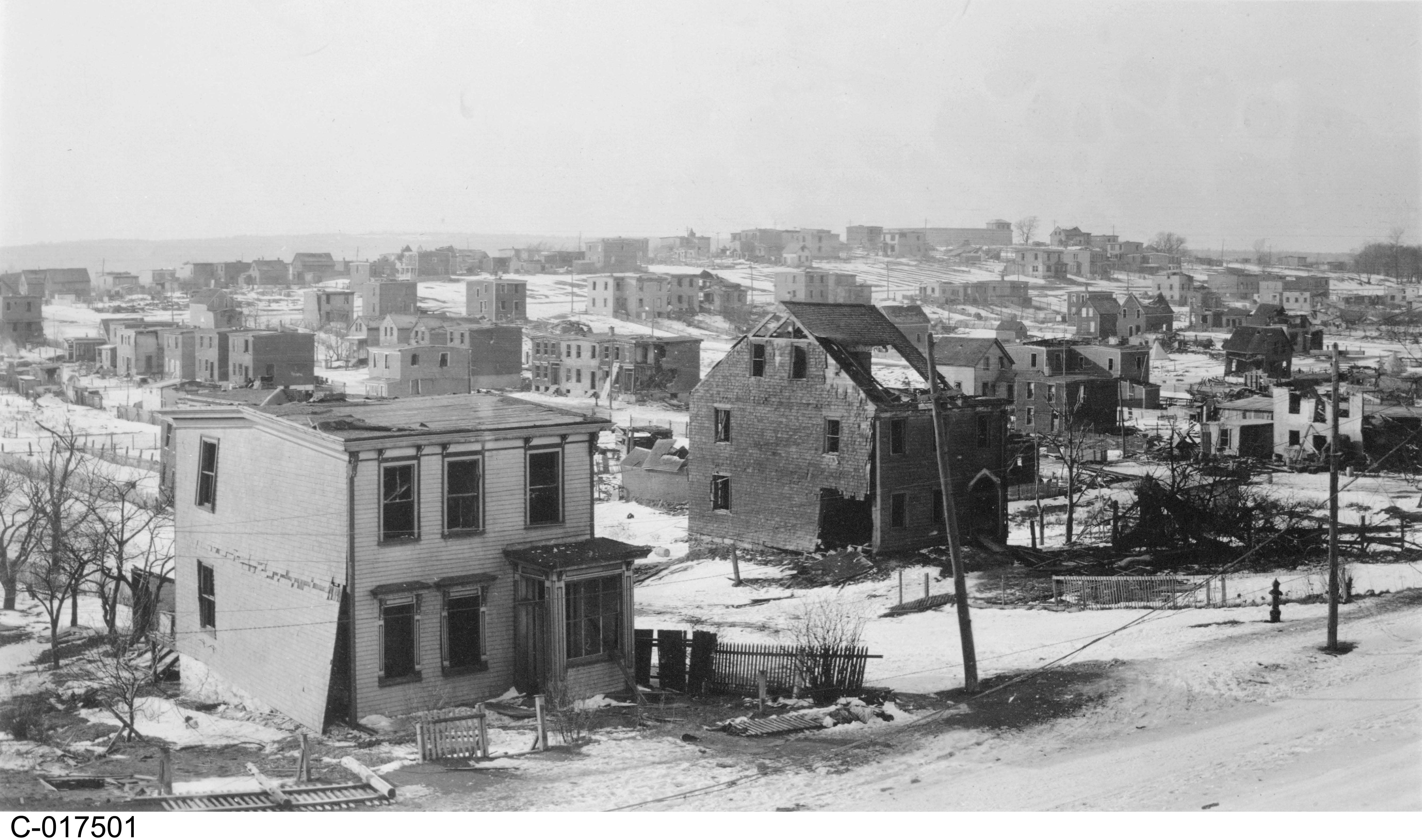 Photographie en noir et blanc – De la neige, des décombres, des arbres calcinés et les ruines de bâtiments sont visibles. Des vitres ont été soufflées loin des maisons. On constate l'omniprésence de cendres.