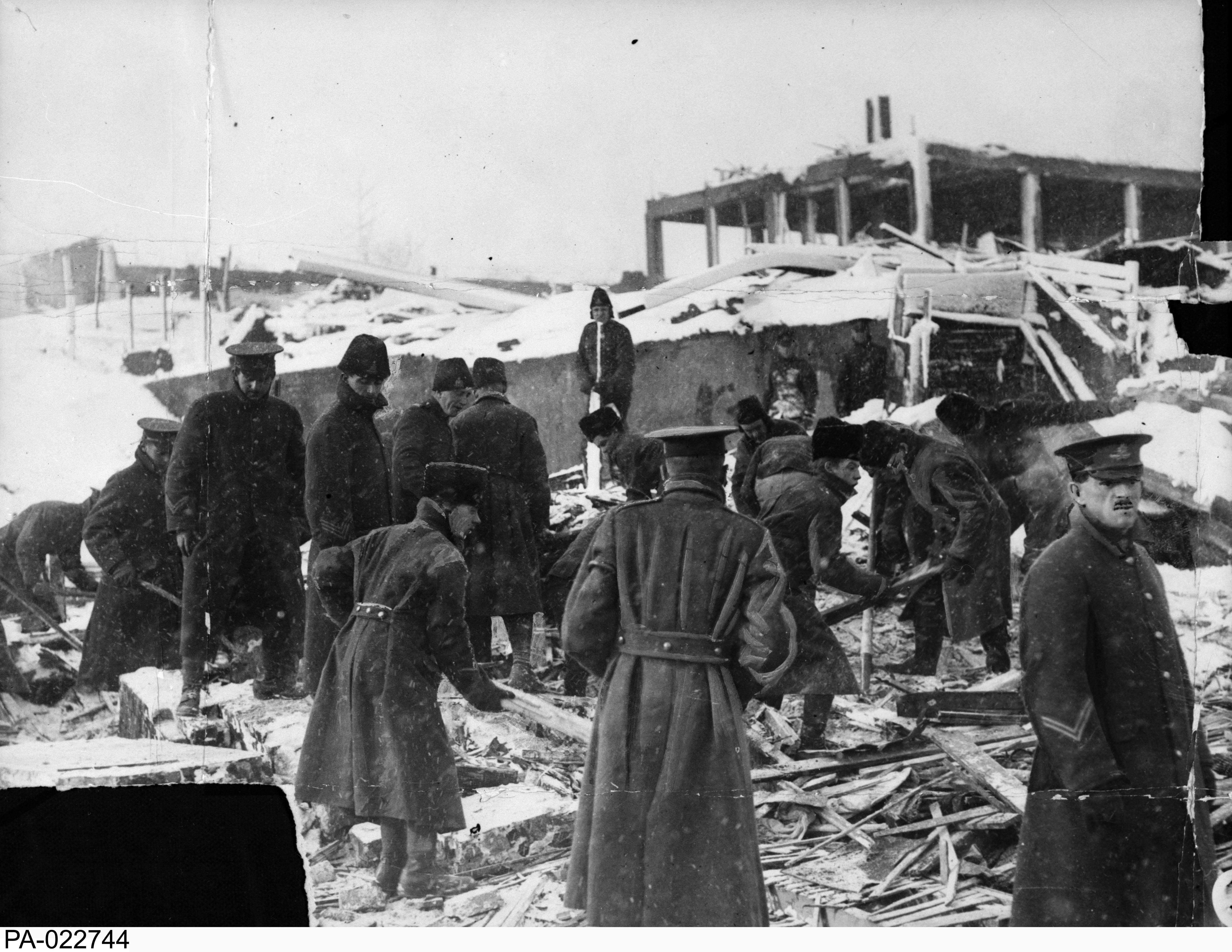 Photographie en noir et blanc – Des soldats fouillent dans les décombres, tentant de retrouver des victimes de l’explosion. Ils portent de longs manteaux d’hiver. Le squelette d’un édifice est visible sur la colline derrière eux.