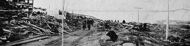 Photographie en noir et blanc – Des survivants avancent le long d’un étroit sentier à travers les débris, loin du rivage. Le squelette d’un immense édifice est visible à gauche.