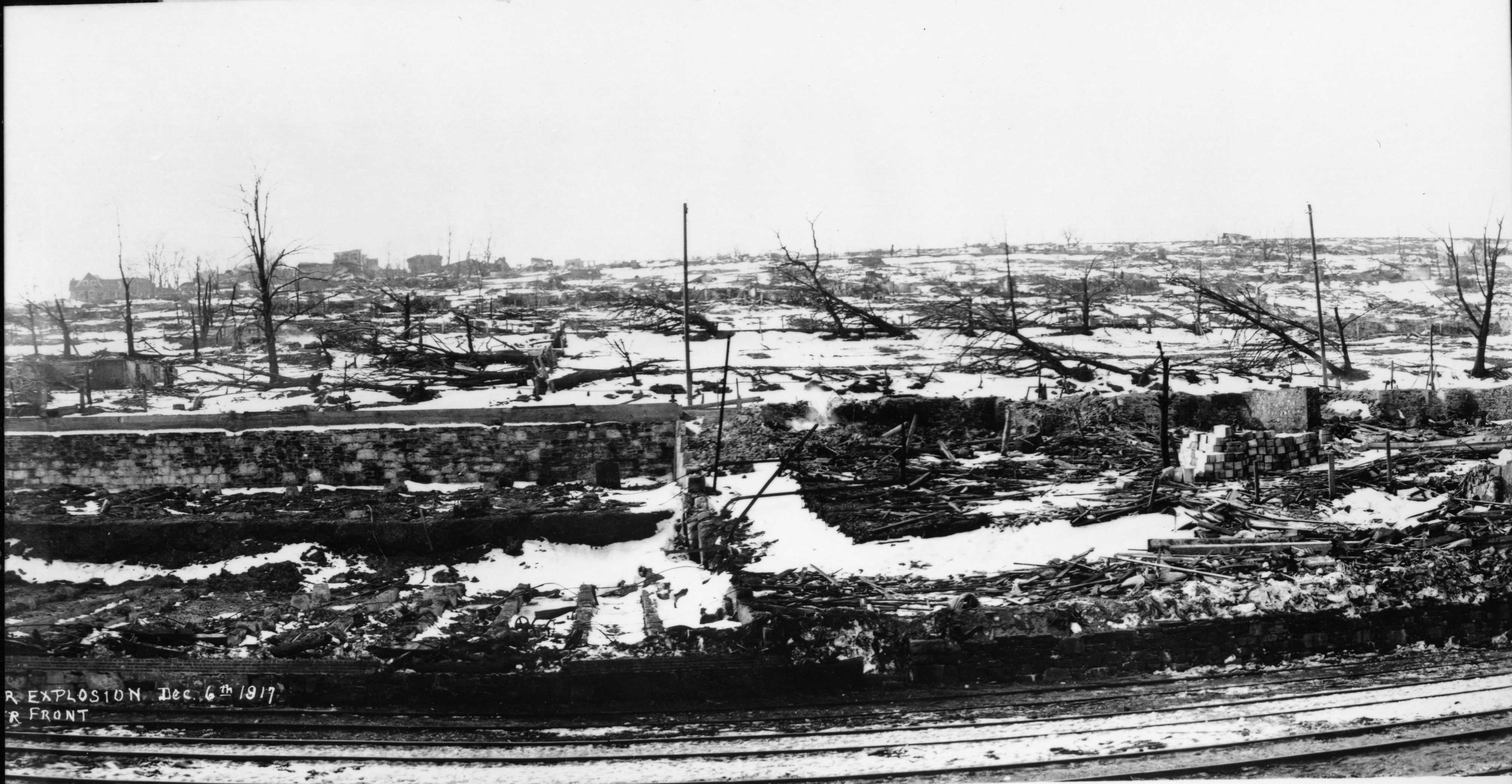 Photographie en noir et blanc – Une voie ferrée au premier plan; quelques structures visibles au loin. On aperçoit la fondation et les décombres d’un bâtiment au centre de la photo, de même que des arbres calcinés et de la neige au sol.