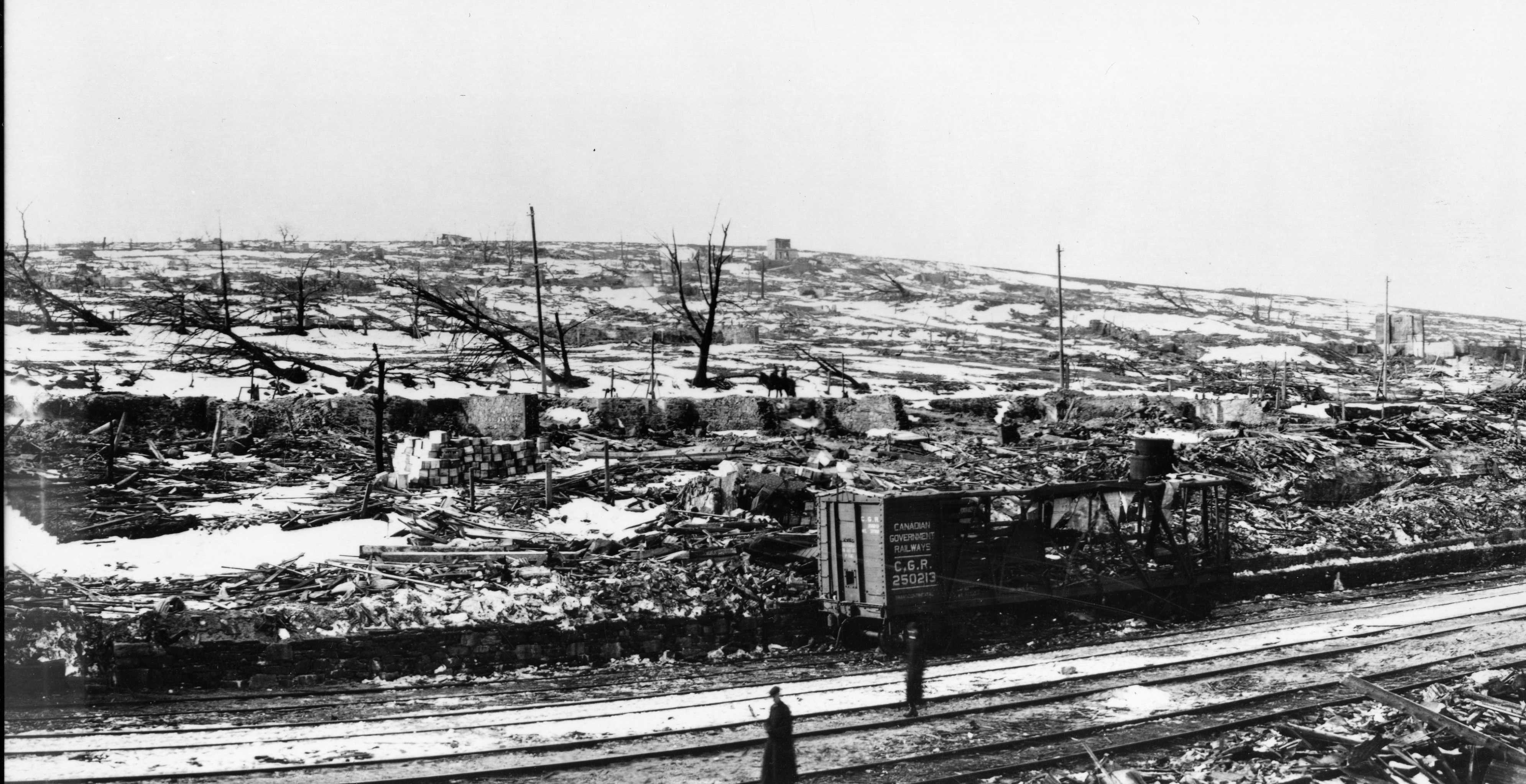 Photographie en noir et blanc – La voie ferrée au premier plan. La neige, les décombres, des arbres calcinés et des ruines de bâtiments composent le paysage. Un détail notable : un véhicule ferroviaire complètement détruit.