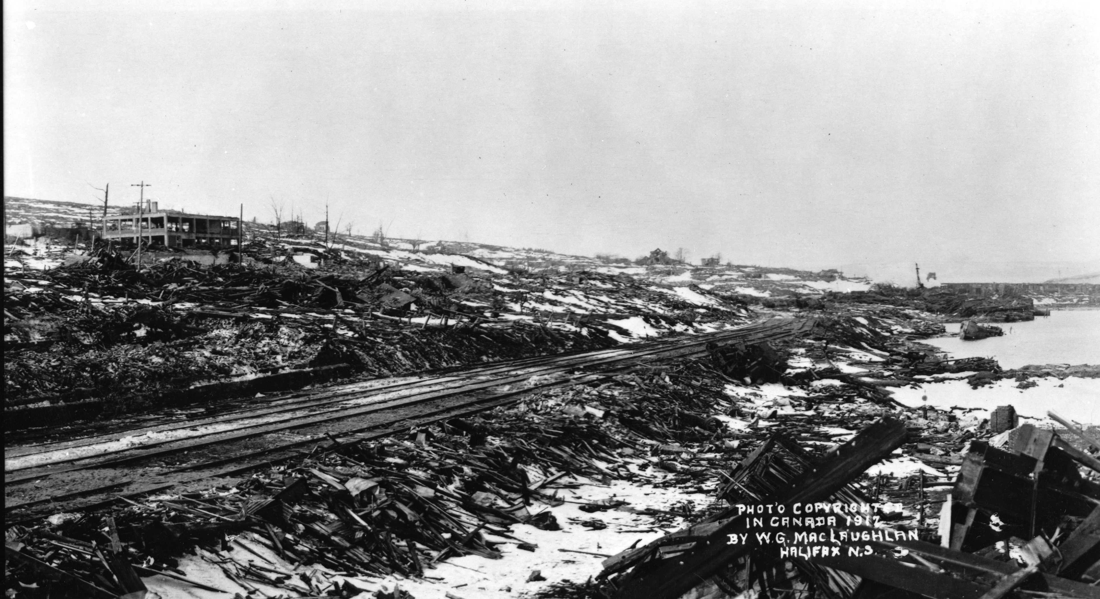 Photographie en noir et blanc – Les voies ferrées se prolongent au loin. On aperçoit un bâtiment soufflé; encore debout, il semble squelettique. Les décombres et les débris sont omniprésents. Les arbres sont calcinés et brisés.