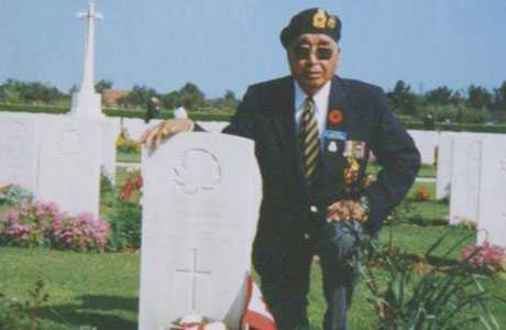 Photographie en couleur – Mark Wolfleg père pose un genou et son bras sur une pierre tombale d'un cimetière militaire. Il porte un veston et une casquette de la Légion.