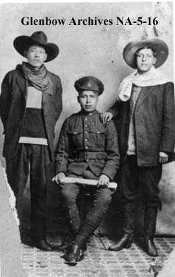 Photographie en noir et blanc – Trois hommes sont photographiés. Deux sont debout, à gauche et à droite de celui assis. Ils portent des vêtements épais et des chapeaux à large rebord, alors que le jeune homme au centre (assis) est en uniforme militaire.