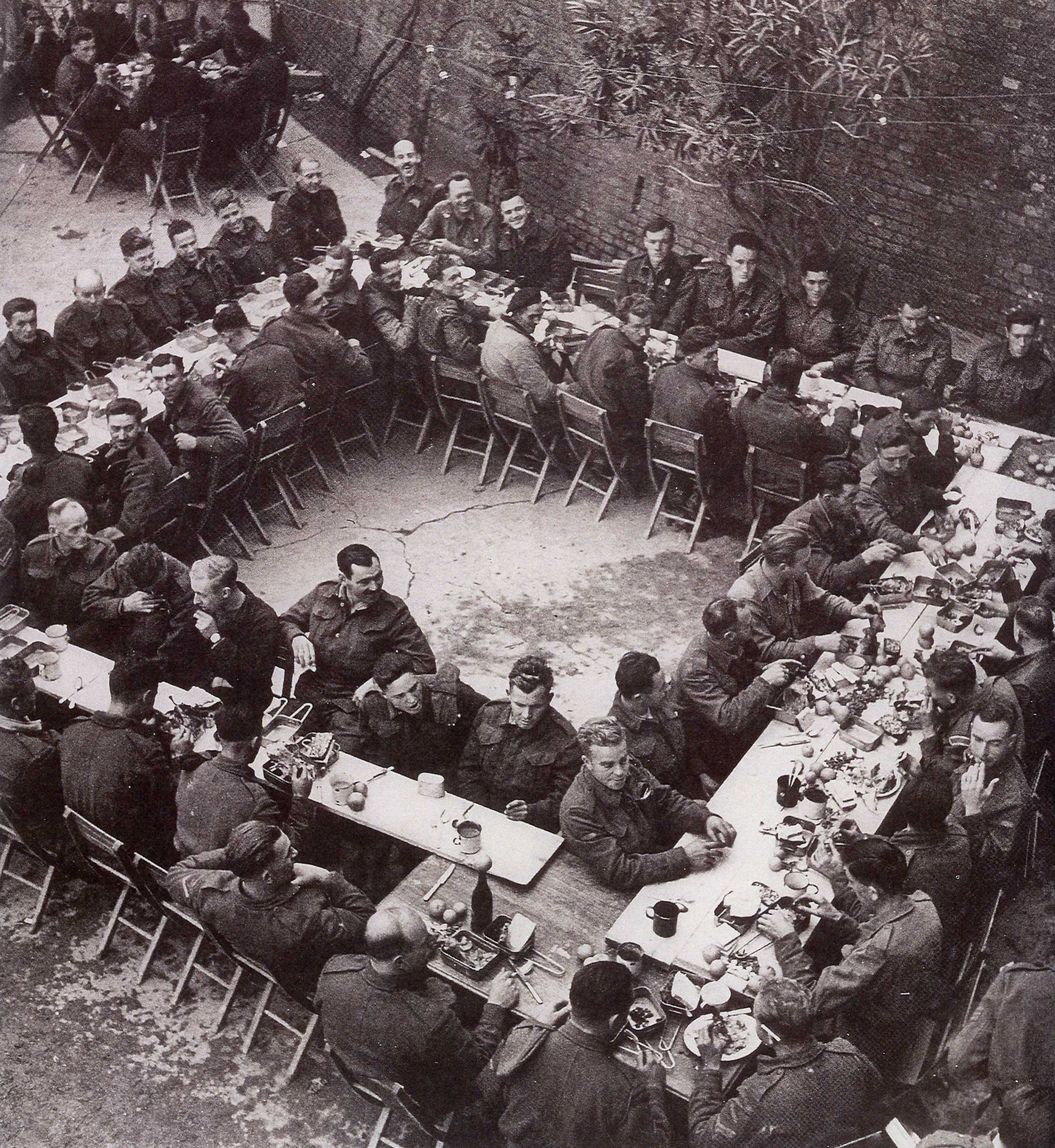Photographie en noir et blanc – Des tables rectangulaires sont disposées de façon à former un grand carré (vue en plongée). Des hommes en uniforme sont assis de chaque côté des tables.