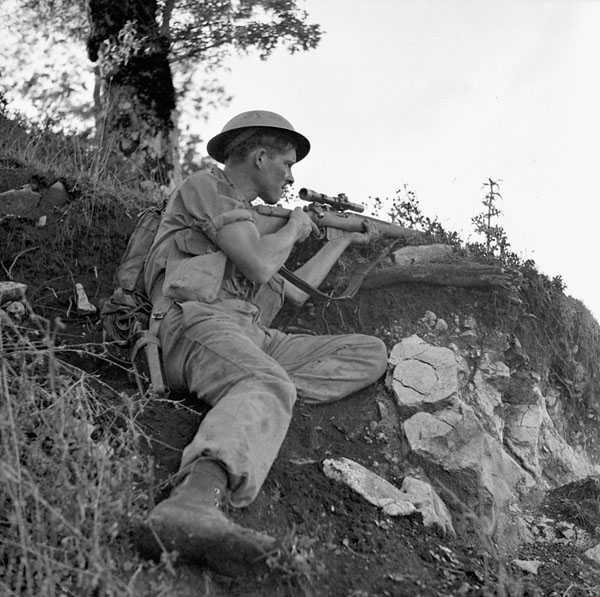 Photographie en noir et blanc – Un soldat canadien est accroupi sous un arbre, derrière un renflement sur un sol rocailleux et broussailleux. Ses manches sont retroussées. Il est prêt à tirer. Ses yeux plissés expriment une concentration élevée.