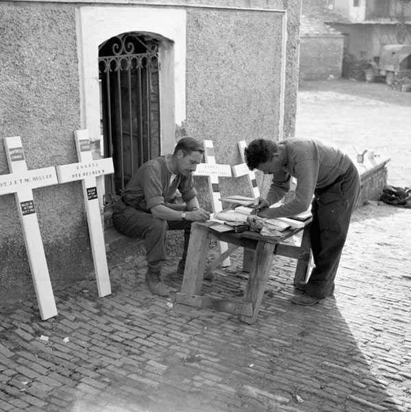 Photographie en noir et blanc – Quatre croix de bois sur lesquelles sont inscrits des noms sont appuyées contre un mur de pierre près d’une clôture métallique. Deux hommes sont penchés sur une petite table en bois en train de préparer d’autres croix.