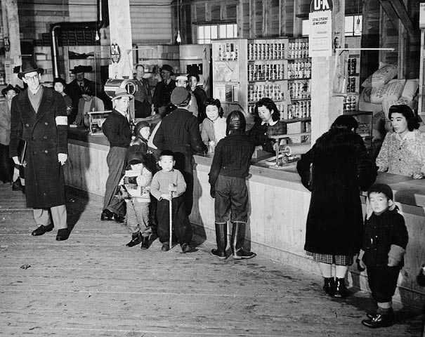 Photographie en noir et blanc – Des gens se trouvent devant et derrière le comptoir d’un magasin. Les tablettes sont remplies de boîtes de conserve et d’autres produits. Les gens conversent de part et d’autre du comptoir.