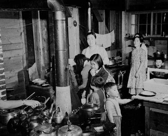 Photographie en noir et blanc – Des Japonaises et leurs enfants sont en train de préparer des aliments dans une cuisine remplie de personnes. On voit un poêle couvert de bouilloires à l’avant-plan et des torchons accrochés à une corde à l’arrière-plan.