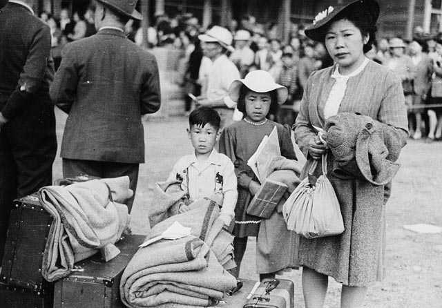 Photographie en noir et blanc – Une femme est debout avec ses deux enfants, endimanchés. On aperçoit tout près un amoncellement de biens personnels et de valises.