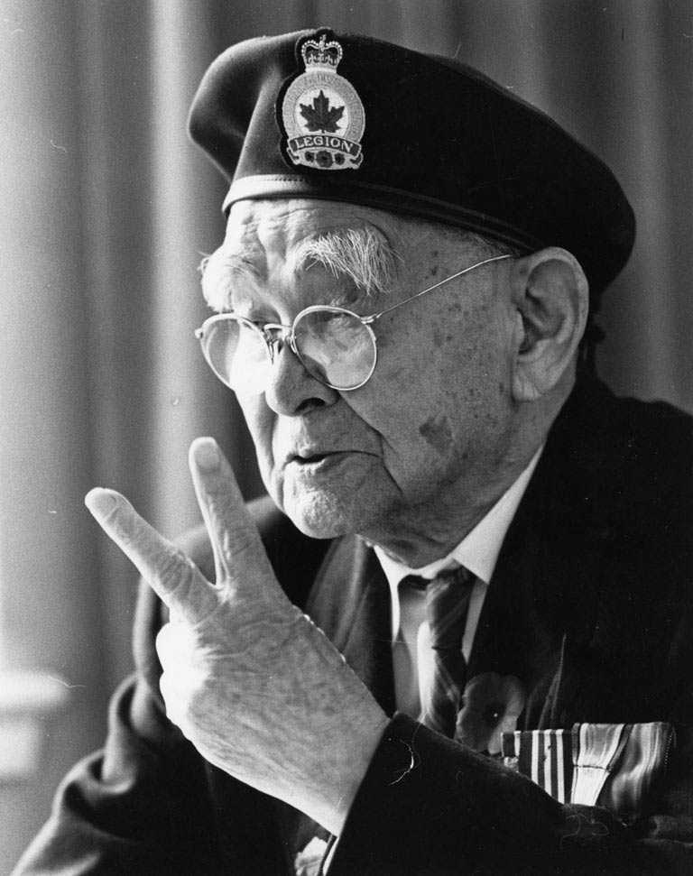 Photographie en noir et blanc – Un Masumi Mitsui âgé fait le signe de la paix. Il porte sa veste de la Légion, des médailles et un coquelicot.