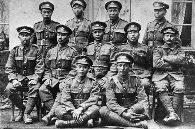 Photographie en noir et blanc – L’image montre onze soldats canadiens d’origine japonaise vêtus d’uniformes militaires et de chapeaux de la Première Guerre mondiale, assis et debout, sur trois rangées, à l’extérieur d’un bâtiment de couleur claire.