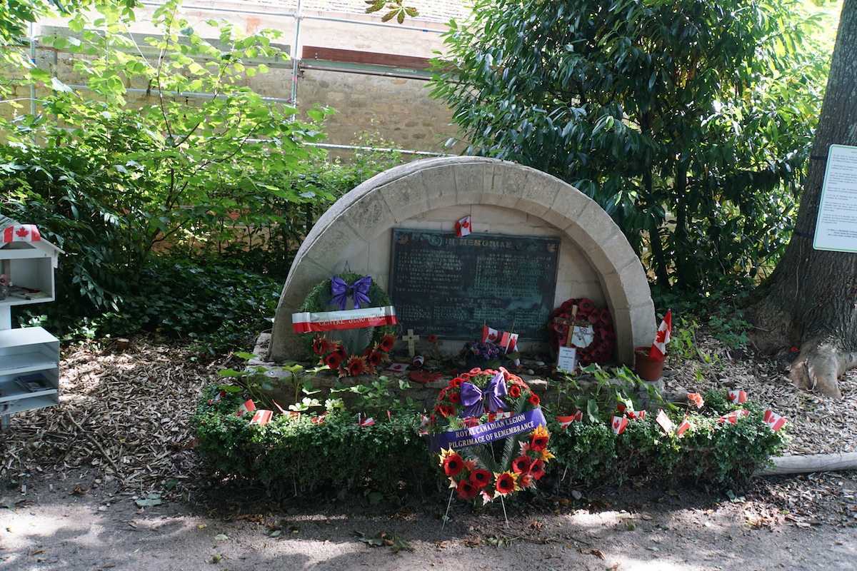 Photographie en couleur – Un petit mémorial en pierre, semi-circulaire, se trouve au milieu d’un jardin luxuriant. Plusieurs couronnes commémoratives ont été déposées à sa base.