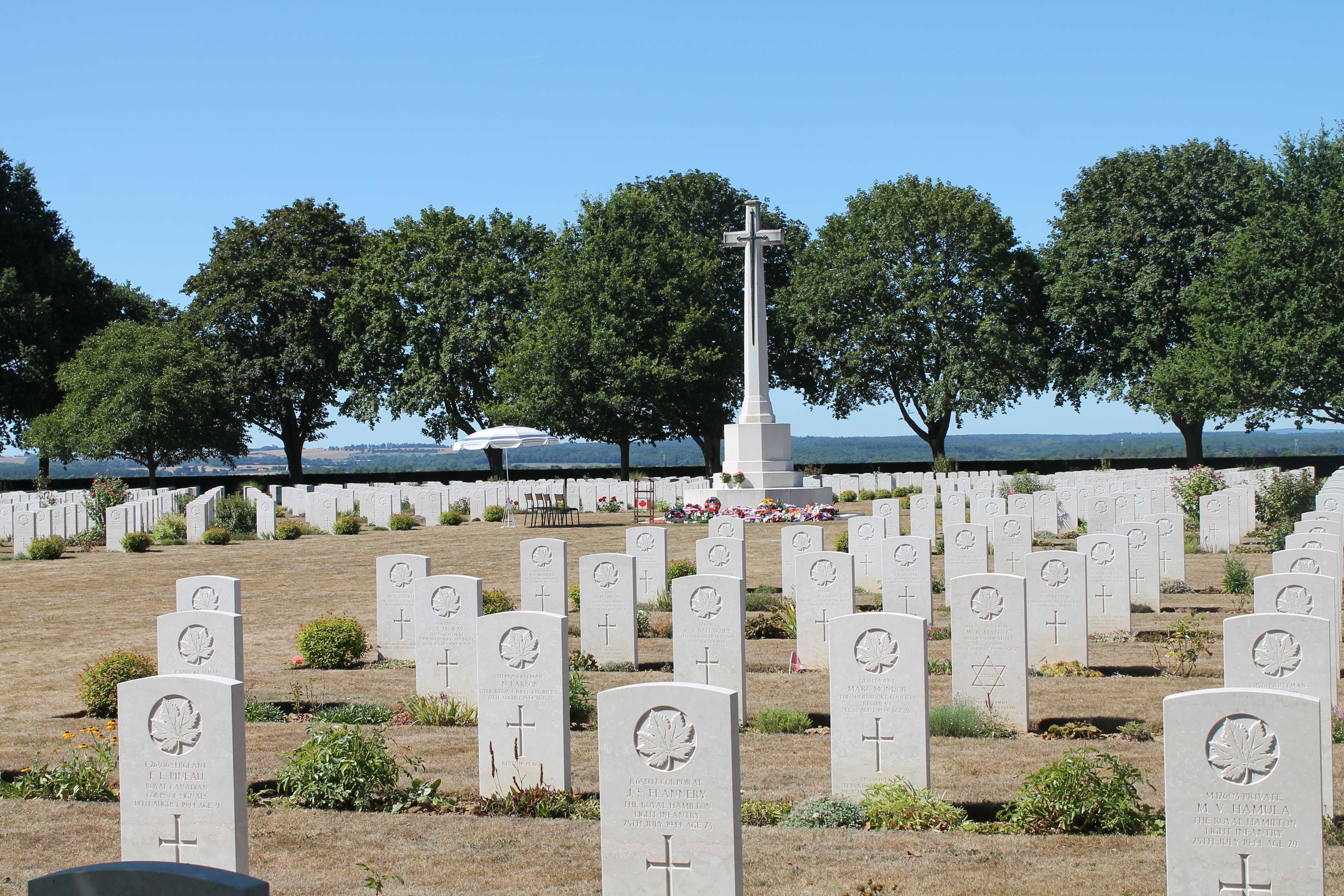 Photographie en couleur – Vue d’un cimetière militaire où se succèdent des pierres tombales blanches uniformes. L’emblématique croix de marbre des sépultures de guerre du Commonwealth est visible au centre. Le podium d’une cérémonie passée est resté.