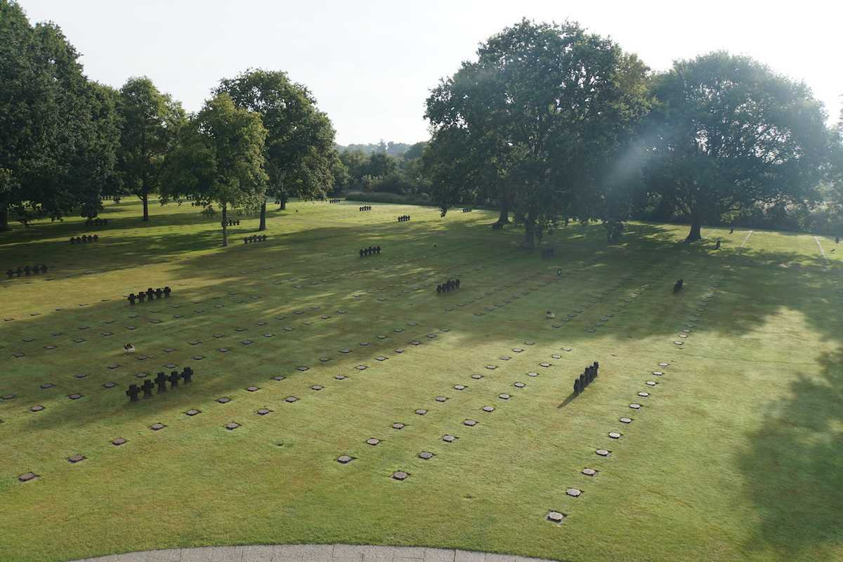 Photographie en couleur – Vue aérienne d’un grand cimetière, semblable à un vaste champ de pelouse. Des pierres plates et des croix en pierre sont disposées à des intervalles réguliers. Les arbres encadrent le paysage.