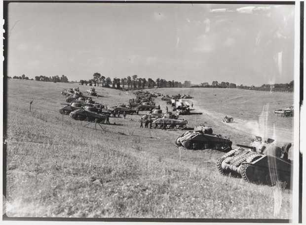 Photographie en noir et blanc – De nombreux chars d’assaut sont stationnés dans le fossé gauche d’une route près d’une zone boisée. Des militaires socialisent autour d’eux.