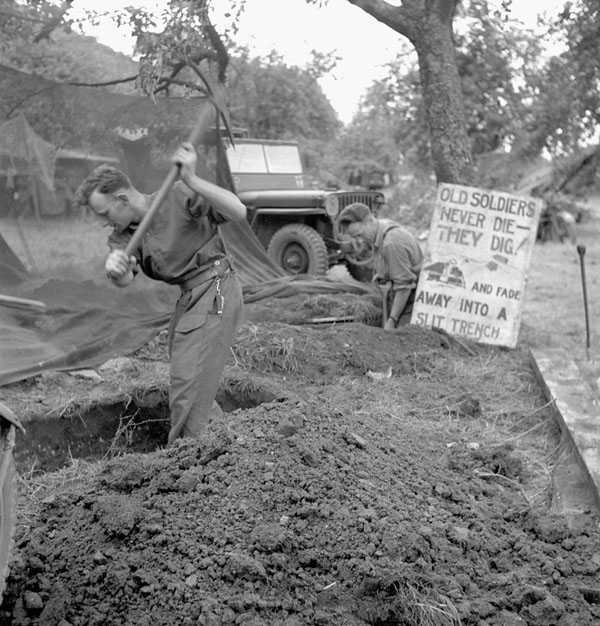 Photographie en noir et blanc – Deux hommes creusant une tranchée étroite dans le sol. On aperçoit des bâches et un tout-terrain militaire à l’arrière-plan, en face d’une zone boisée.