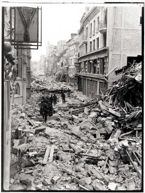Photographie en noir et blanc – Des troupes canadiennes en rangée descendent le long d’une route étroite à Caen. Les bâtiments de part et d’autre montrent de nombreux dommages. Les rues sont complètement couvertes de gros morceaux de gravats.