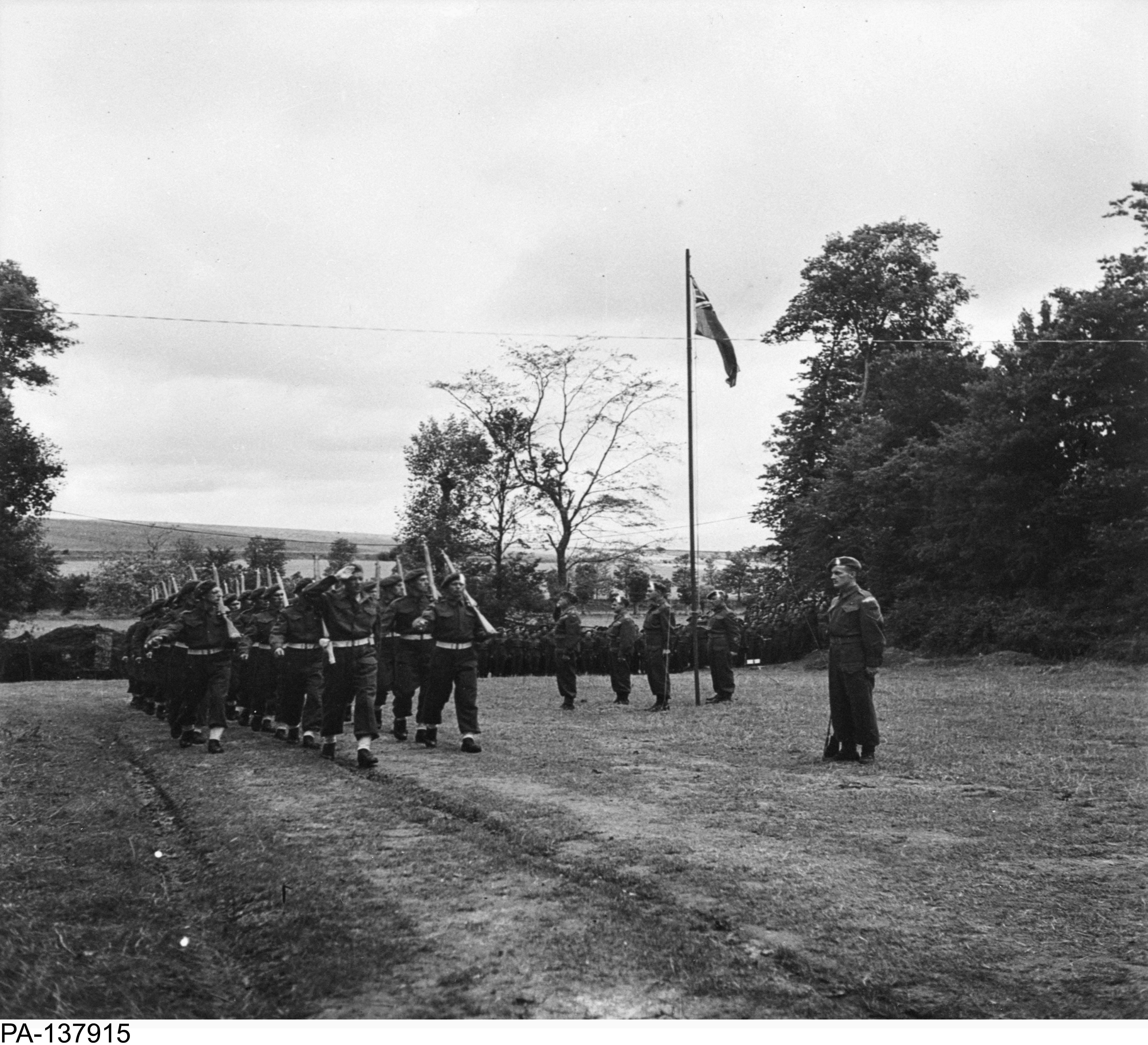 Photographie en noir et blanc – Trois rangées de soldats défilent derrière un drapeau Red Ensign et des officiers supérieurs. Tous adressent un salut. Ils sont dehors, dans un champ gazonné et entouré d’arbres.