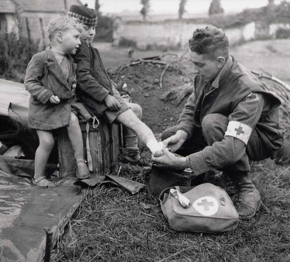 Photographie en noir et blanc – Un soldat (avec un brassard médical) bande la cheville d’un garçon sous le regard attentif d’un autre enfant.